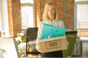 Woman holding a cardboard box labeled "Fired," standing in an office after losing her job.