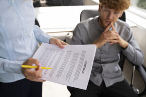 Close-up of a person reviewing a contract with a pen while another person listens attentively in an office.