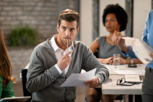 Thoughtful businessman holding a pencil, attending a meeting with colleagues in a workplace setting.