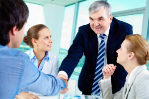 Senior executive in a business suit shaking hands with a colleague in a meeting, with two other professionals looking on.