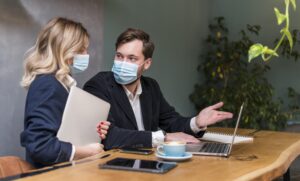 Two colleagues in business attire wearing masks, discussing work over laptops and coffee in a workplace setting.